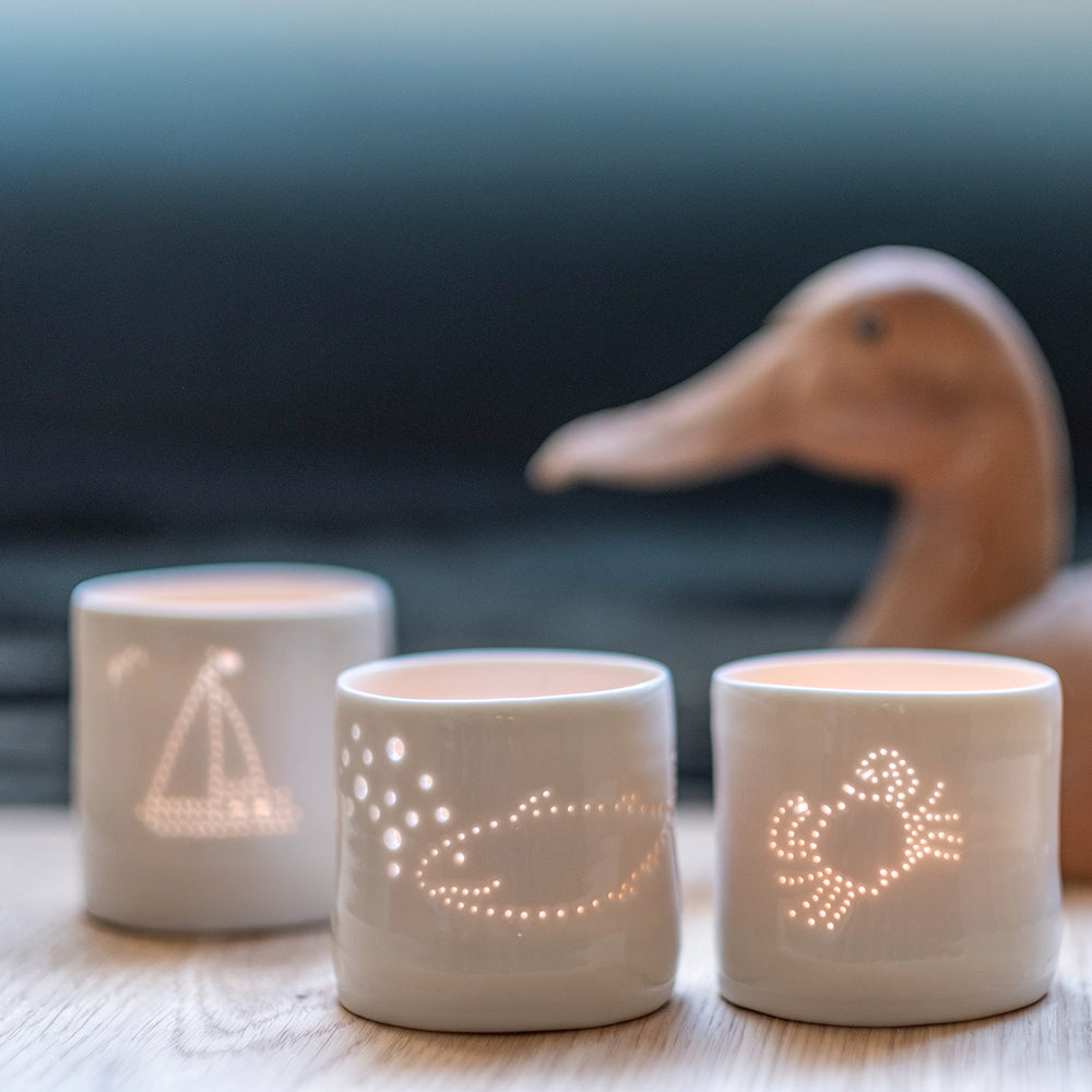 Three porcelain tealight holders, with a crab design, a whale, and a sailing boat. The designs are pierced into it with tiny holes that let the light shine through. These ceramic candle votives are perfect for seaside holidays.
