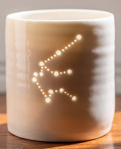 A ceramic porcelain tealight holder, with the Aquarius constellation pierced into it with tiny holes, allowing candlelight to twinkle through. The tealight holder has natural glossy glaze and is almost translucent.