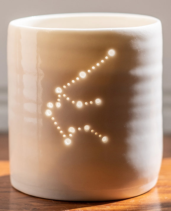 A ceramic porcelain tealight holder, with the Aquarius constellation pierced into it with tiny holes, allowing candlelight to twinkle through. The tealight holder has natural glossy glaze and is almost translucent.