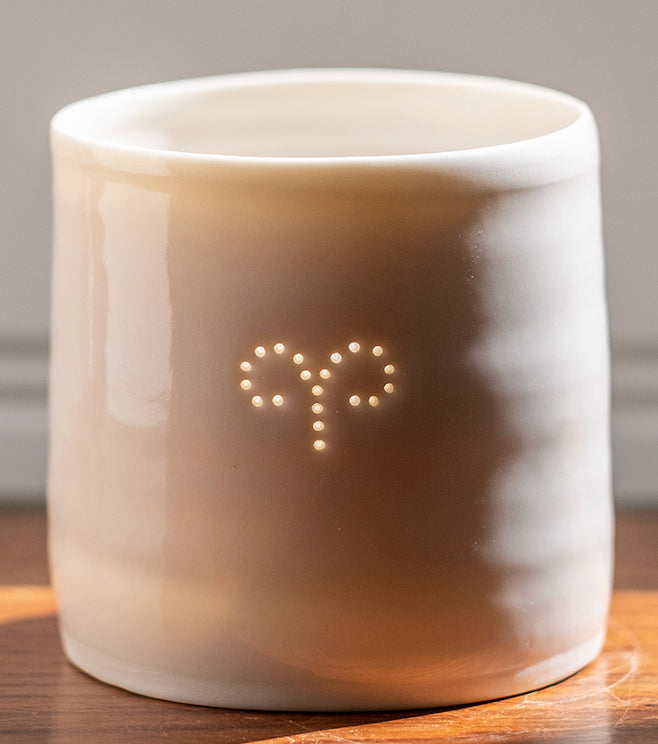 A ceramic porcelain tealight holder, with the Aries astrological symbol pierced into it with tiny holes, allowing candlelight to twinkle through. The tealight holder has natural glossy glaze and is almost translucent. Relaxing homeware.