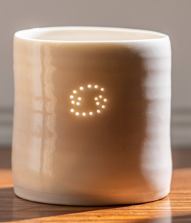 A ceramic porcelain tealight holder, with the Cancer astrological symbol pierced into it with tiny holes, allowing candlelight to twinkle through. Natural glossy glaze, almost translucent. Horoscope, astrology, zodiac.
