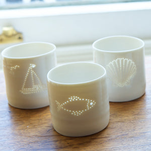 Part of the seaside collection, this porcelain tealight holder features a fish design. This ceramic candle holder is pierced with tiny holes to create a natural glossy glow. Here with seashells and sailing boats.